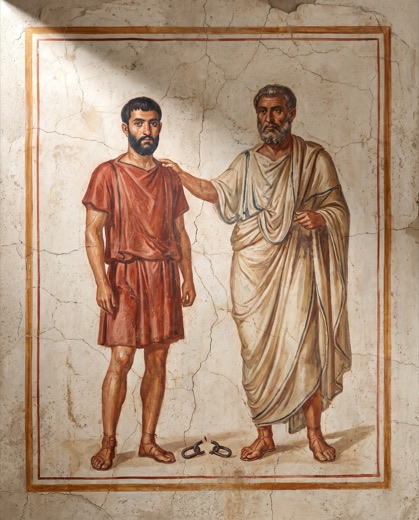 El patrono pone la mano en el hombro del liberto ante una losa de piedra, con el eslabón partido en el suelo.
